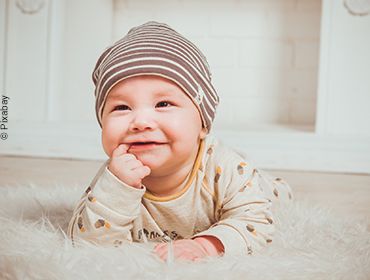 L&auml;chelndes Baby liegt auf dem Bauch eines Teppichs mit erhobenem Kopf und einem Finger im Mund